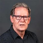 A middle-aged man with short gray hair, wearing glasses and a black polka dot shirt, looking at the camera against a plain gray background.