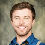 A man with short hair and a beard, wearing a blue shirt, tie, and suspenders, smiles at the camera against a mottled backdrop.