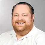 A man with a beard and mustache smiles at the camera, wearing a white collared shirt against a plain gray background.