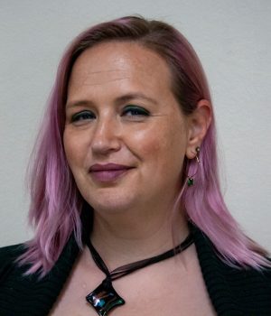 A person with shoulder-length pink hair, wearing a black top, necklace, and earrings, stands in front of a plain light-colored wall, looking at the camera with a neutral expression.